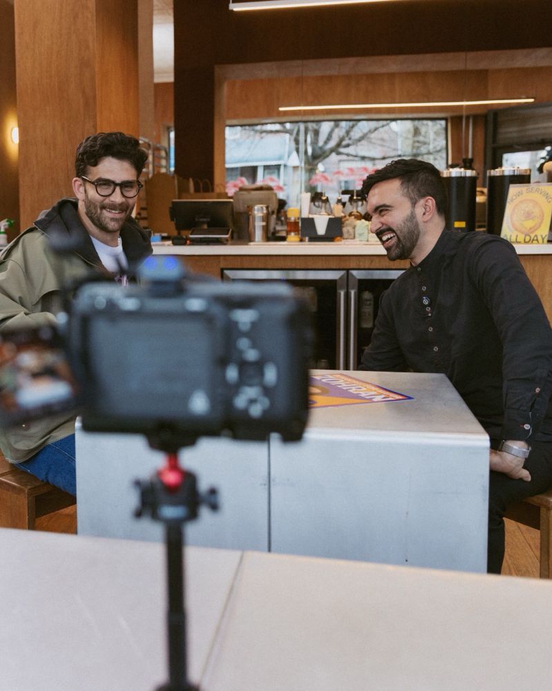 Hasan and Zohran sitting at a table, both smiling, in Little Flower Cafe, with a camera pointed at them in the foreground.