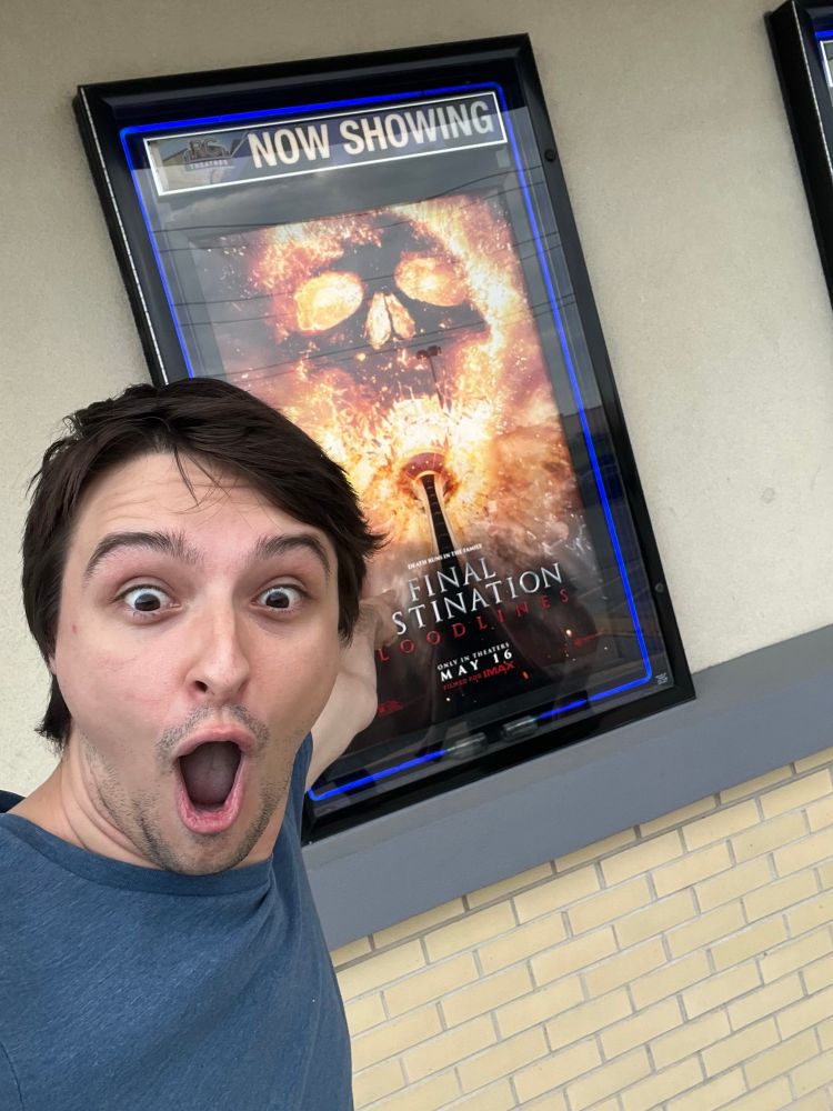 Picture of Trent doing the soyjack point with mouth a gape pointing at a poster for final destination bloodlines