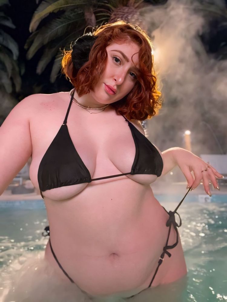A red head with short hair and black earmuffs wearing a small black bikini that she is spilling out of and staring at the camera seductively in a hot tub. Steam rises around her as she pulls on the string of her bottoms with her left hand. Palm trees can be seen through the haze behind her. 