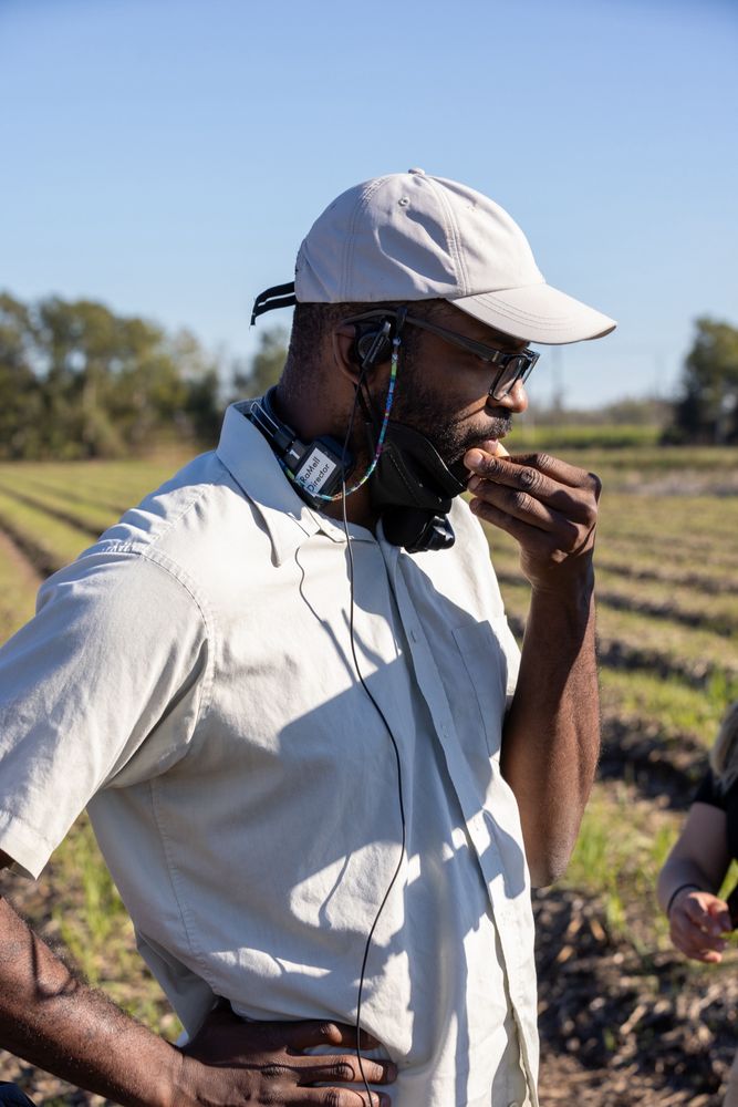 RaMell Ross wears a white shirt and ballcap while holding his fingers up to his mouth in a field while directing Nickel Boys. 