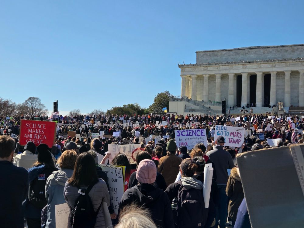 Picture shows The Lincoln Memorial in the top right corner. The sky is blue, and the bottom half is completely filled with a crowd of people and 3-4 signs about Science. One sign in the bottom left has a red background and says "SCIENCE MAKES AMERICA GREAT" in white letters. 