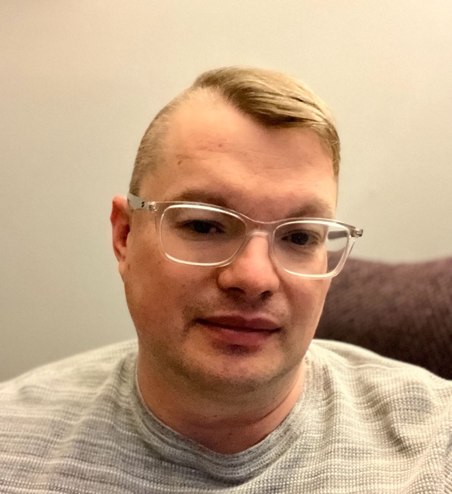 White male wearing clear plastic glasses, straight brown hair swept to one side, the other side shaved close to his skin 