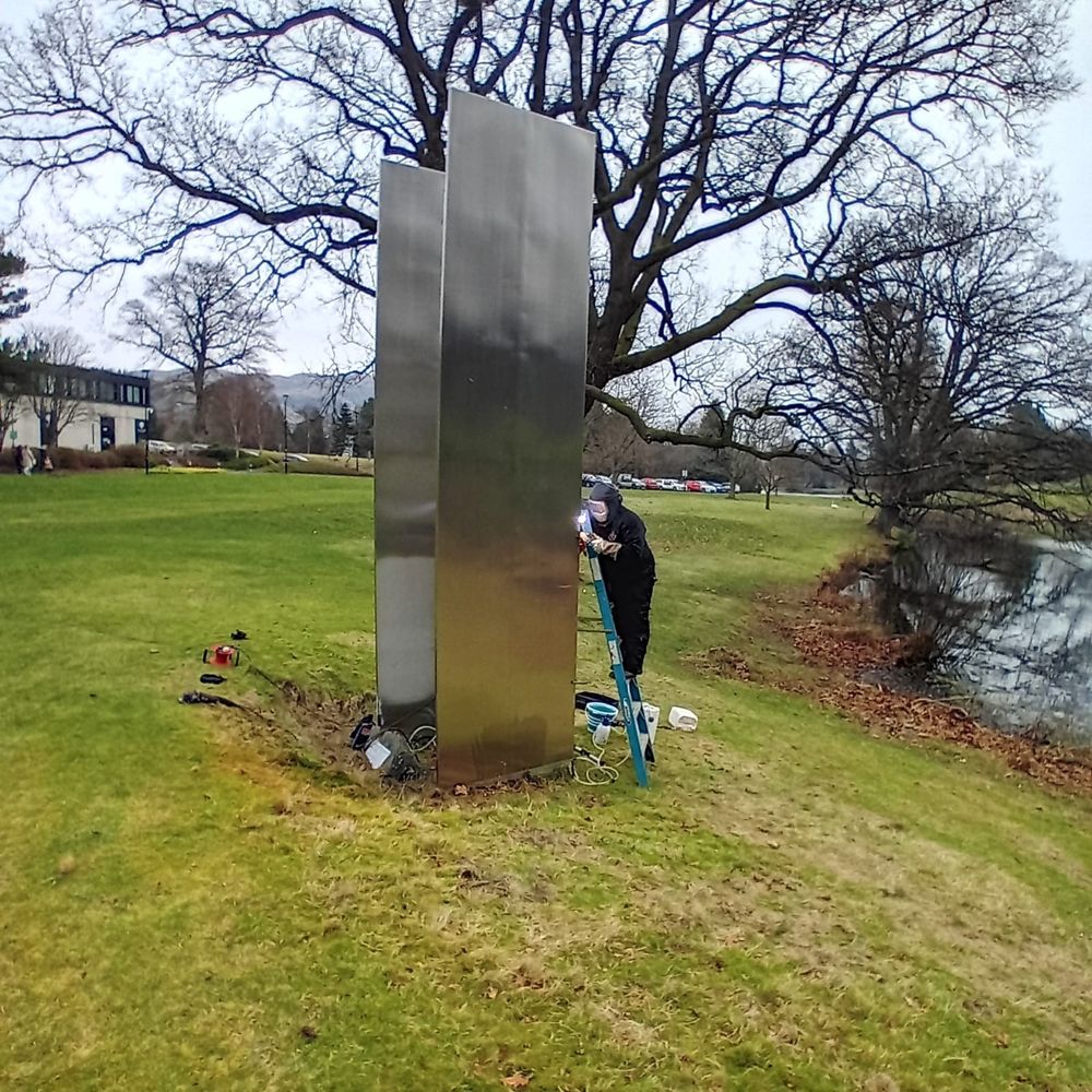 Two upright rectangular sculptures facing each other on a grass lawn with a loch and buildings and trees in the background. There is a person on a ladder, wearing PPE and using a tool on the sculpture. The two sculptures are now a shiny steel 