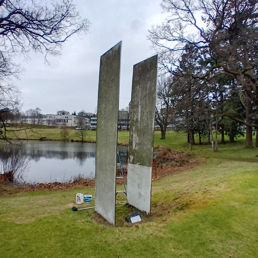 Two upright rectangular sculptures facing each other on a grass lawn with a loch and buildings in the background and some trees at the edges of the image. The sculptures look grainy,mossy and dirty, with some of the dirt been removed on the sculpture to the right. 
