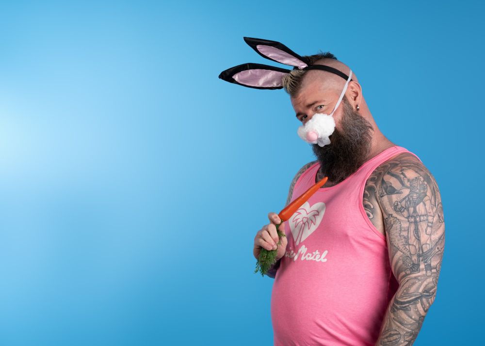 A bearded, tattooed man wearing bunny ears and a bunny nose holding a plastic carrot in front of a blue backdrop. 