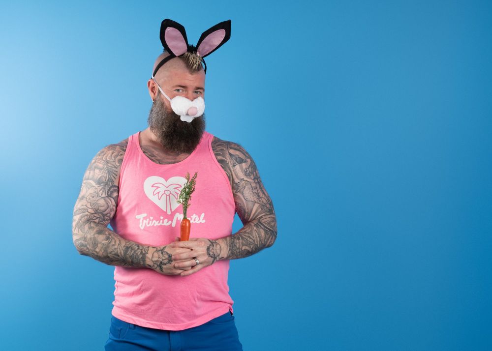 A bearded, tattooed man wearing bunny ears and a bunny nose holding a plastic carrot in front of a blue backdrop. 