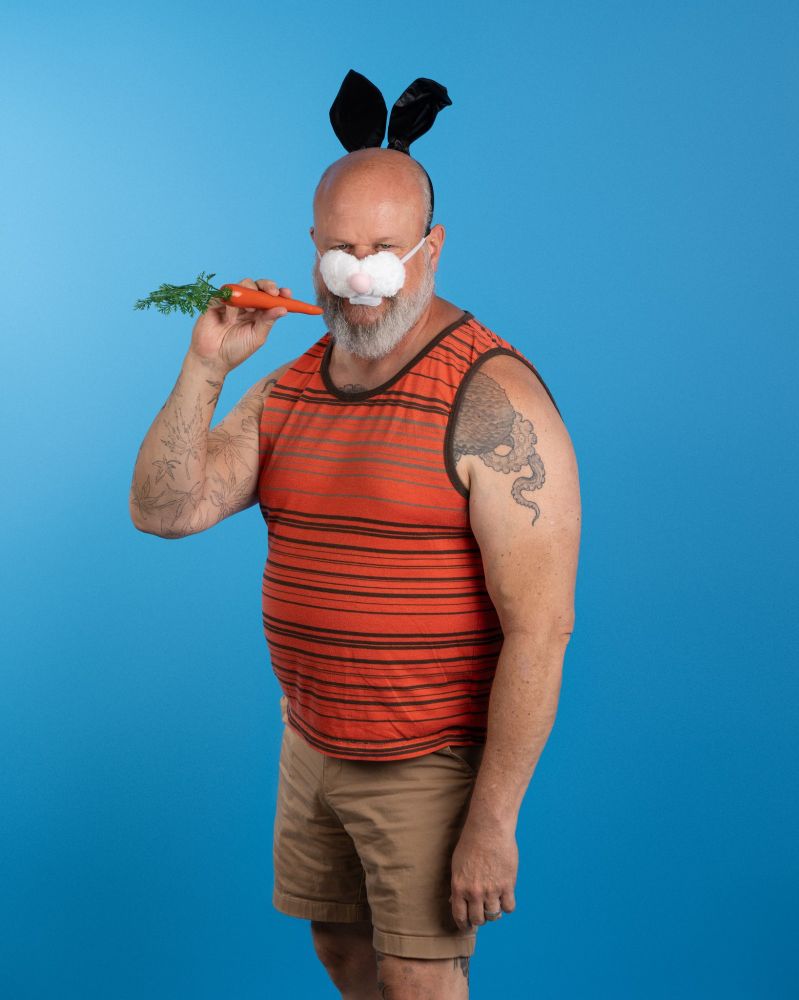 Eric, wearing bunny ears and a bunny nose and holding a plastic carrot in front of a blue backdrop 