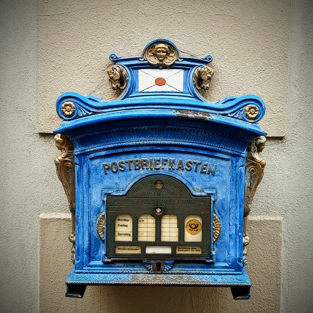 Blauer,verschnörkelter,alter Postbriefkasten an einer hellen Wand 