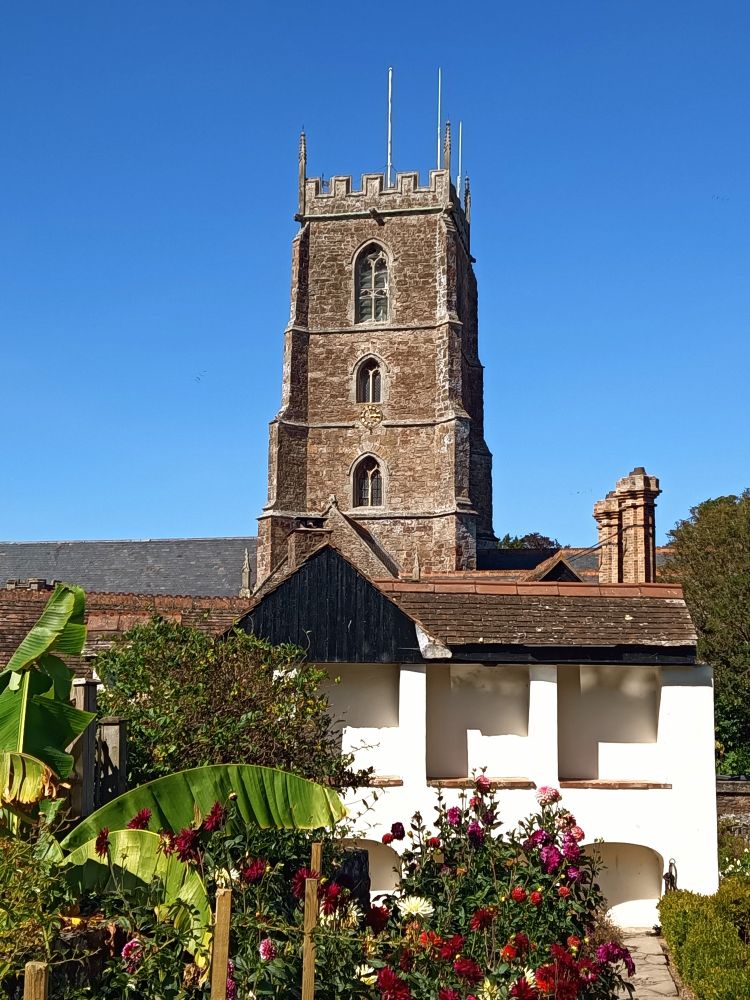Dicker viereckiger Kirchturm aus Naturstein,davor eine kleine Gartenanlage mit Dahlien und einer Palme, weißes Gebäude mit Ziegeldach unterhalb des Kirchturms 