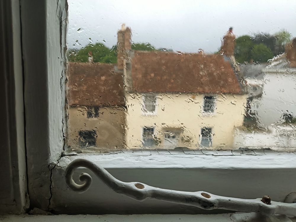 Teil einer Fensterscheibe, Regen auf ihr lässt die Häuser auf der anderen Straßenseite verschwimmen.weisser Holzfensterrahmen und Teil eines Fensterverschlusses