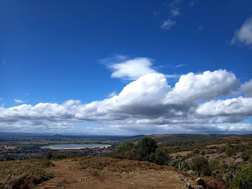 Blick über die Landschaft vom Cliff aus.Dicke weiße Wolken am blauen Himmel. Hügel, Büsche, Bäume,die Stadt Cheddar und das Cheddar Reservoir in der Ferne 