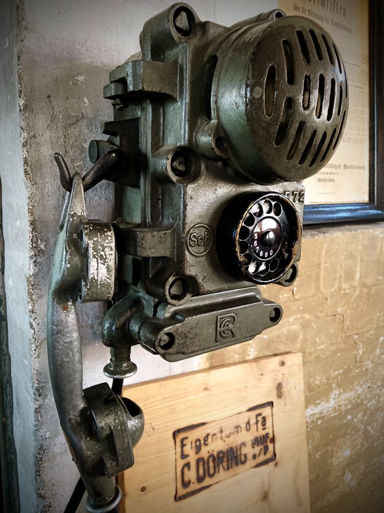 Telefon in einer Werkhalle, groß,klobig,aus Metall, schwarz mit kleiner Wählscheibe unten, Hörer links an einem Haken eingehängt 