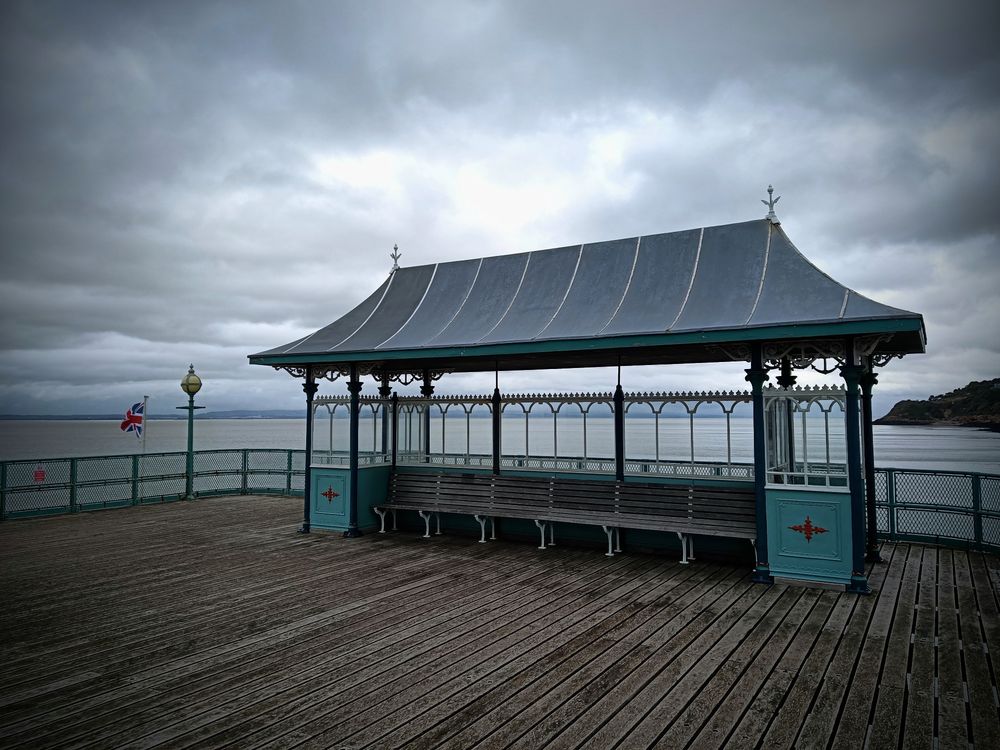 Kleiner Pavillon mit Sitzbänken auf dem Pier, Holzplanken, Geländer ringsherum,eine britische Flagge weht, grauer Himmel 