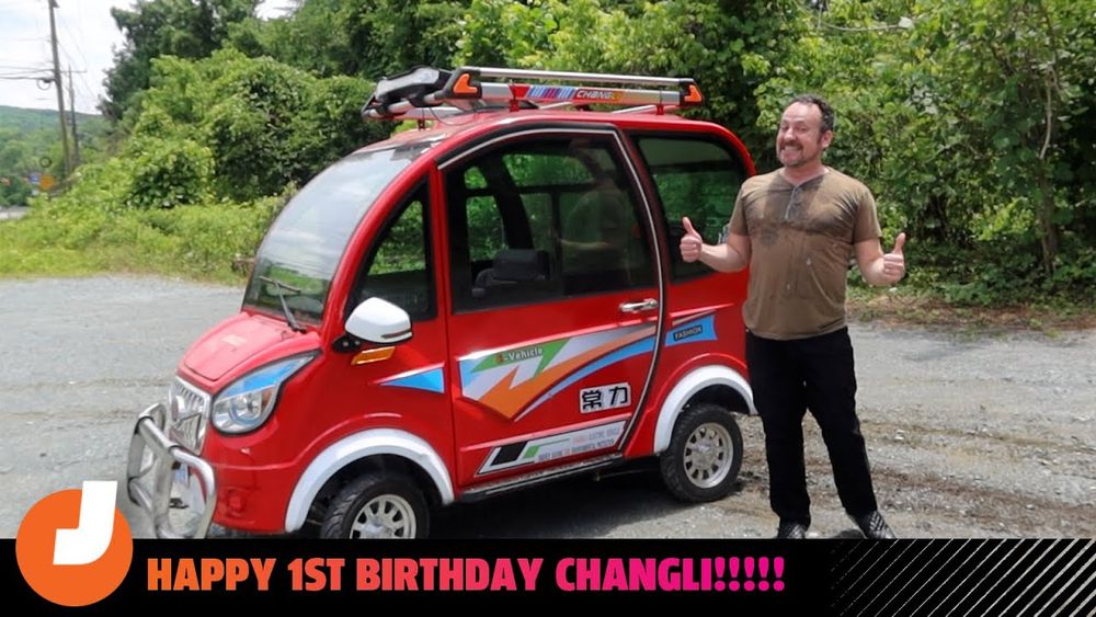 A man standing in front of an automobile of extreme elegance and utility, or rather a cheap Chinese EV with a bulging cabin.