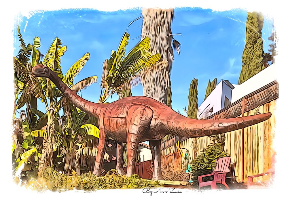 Large dinosaur sculpture in a tropical garden. This scene looks stylized, like a painting or digital art. The dinosaur resembles a sauropod, with a long neck and tail, standing among tall palm trees and banana plants. Bright blue sky forms the background, and the area looks fenced with a wooden structure. There are two pink chairs under the dinosaur’s tail, and the overall atmosphere is vibrant and whimsical. At the bottom, the text reads: By Ann Zeise.
