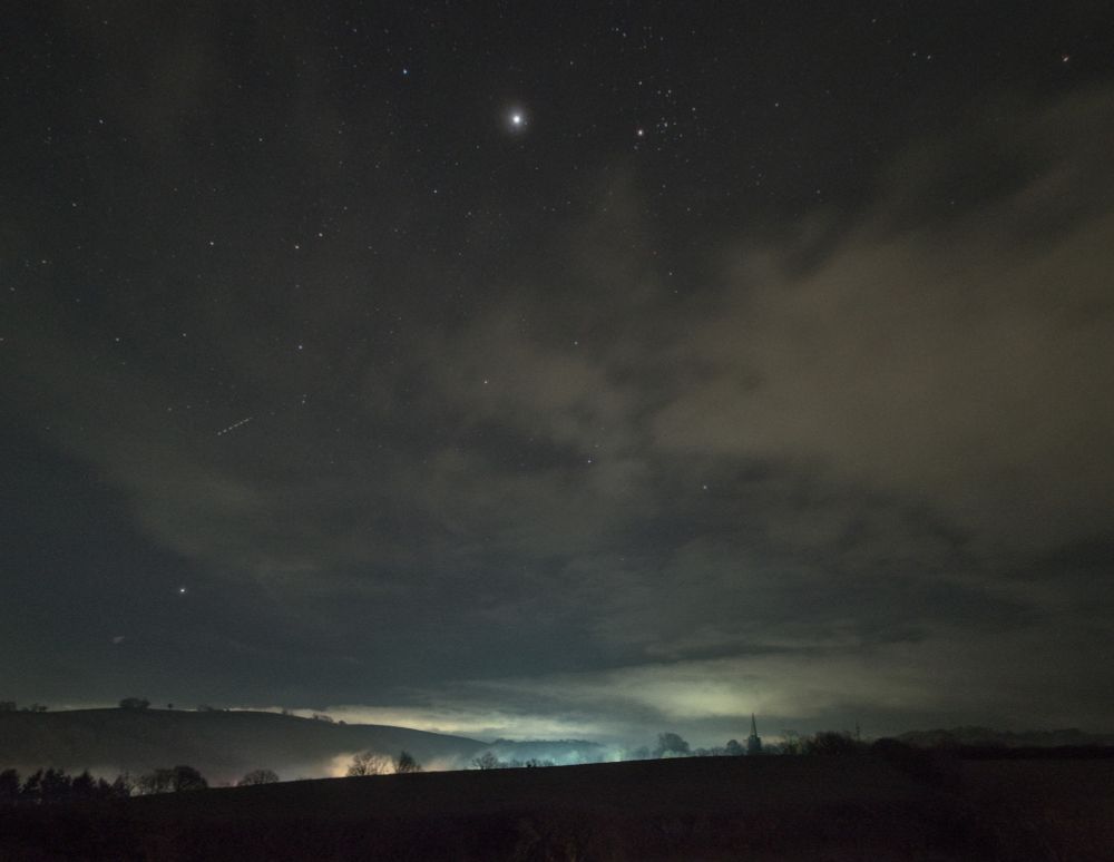 Night photograph. Orion centred, immersed in cloud. Jupiter shining extremely brightly, showing as a bright smudge due to haze and glare. The V shape of Taurus in a relatively clear patch of sky to the right. The lower part of the image is the ground, dark hills, mist in the valley, a church spire breaks the horizon on the centre right. 