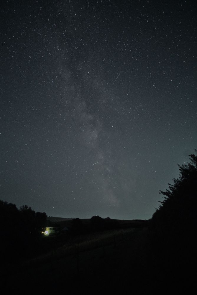 Starry, cloudless, night scene photo. Field in the foreground, with fence posts going to a vanishing point on the right. Single house light shines brightly on the far side of the field. Hedgerow high on the right. Milky way starts in the top left, and finishes just off to the right of centre near the horizon. The image has been processed to appear reasonably ‘natural’ so the cloudiness of the Milky Way is whitish. There are two planes that have drawn short lines in the sky during the exposure. The rest of the sky is densely packed with stars, the ‘teapot’ asterism in Sagittarius is low in the centre, and the ‘Altair Family’ appears quite clearly near the top left. 