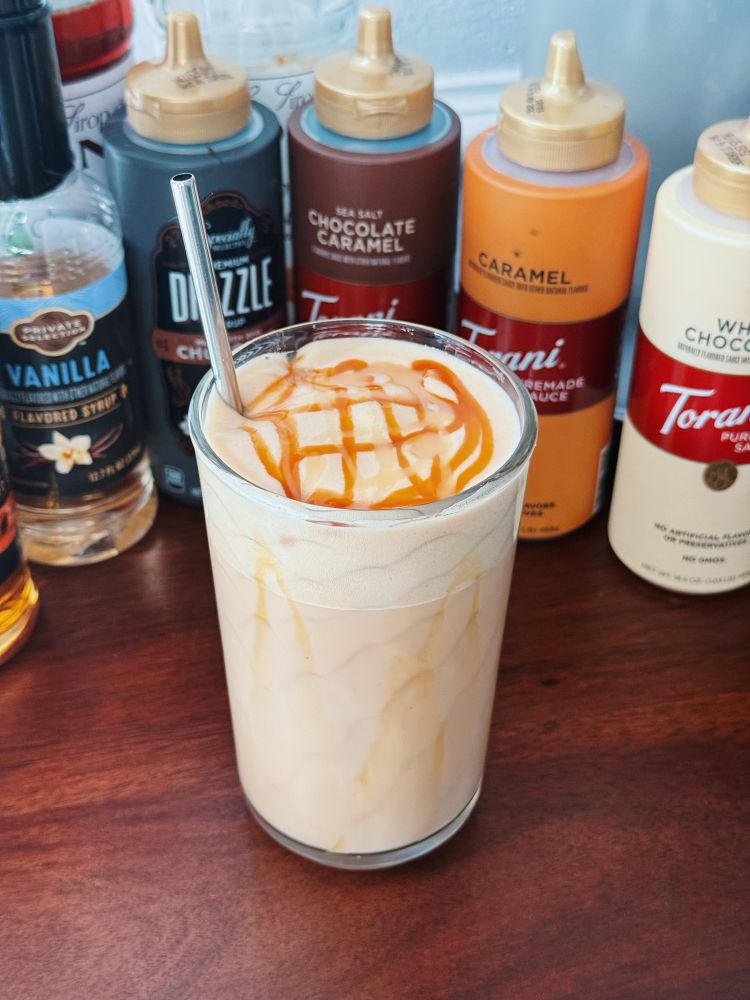 A caramel and white chocolate iced latte in a glass, topped with caramel macchiato cold foam and drizzled with caramel and white chocolate sauces at the top, it also has a stainless steel straw. Surrounded it is the various coffee sauces and syrups at my coffee bar. 
