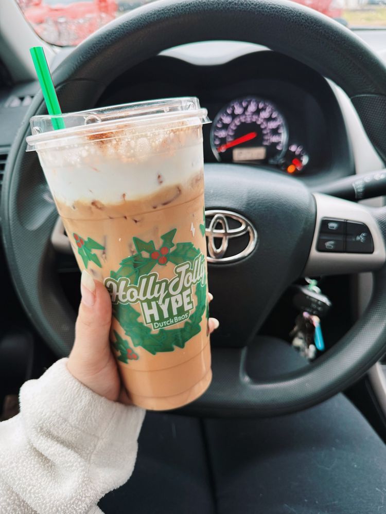 A large iced frosted gingerbread latte with oat milk from Dutch Bros being held in front of a Toyota steering wheel. You can also see on my dashboard that my gas light is on. 