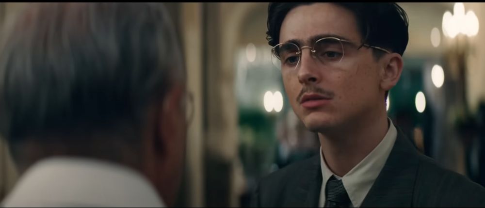 A screencap from a trailer, showing Timothée Chalamet with glasses and a thin moustache, in a period piece of some sort. He's talking to a man in the foreground.