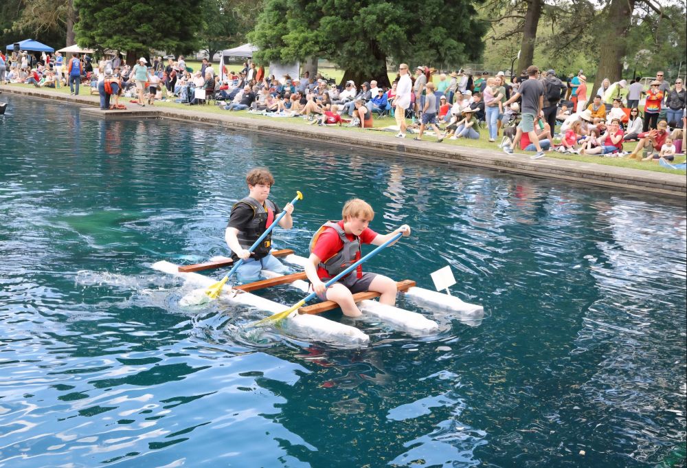 2 kids paddling a homemade boat with spectators watching