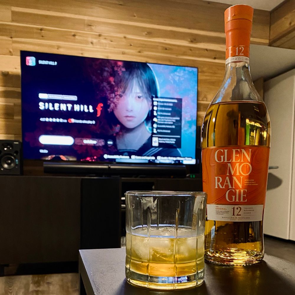 Square photo of a bottle of Glenmorangie 12 year scotch with a glass of said scotch next to it on a black table surface. In the background a large TV can be seen with SILENT HILL f downloading onto a PS5 on the screen.