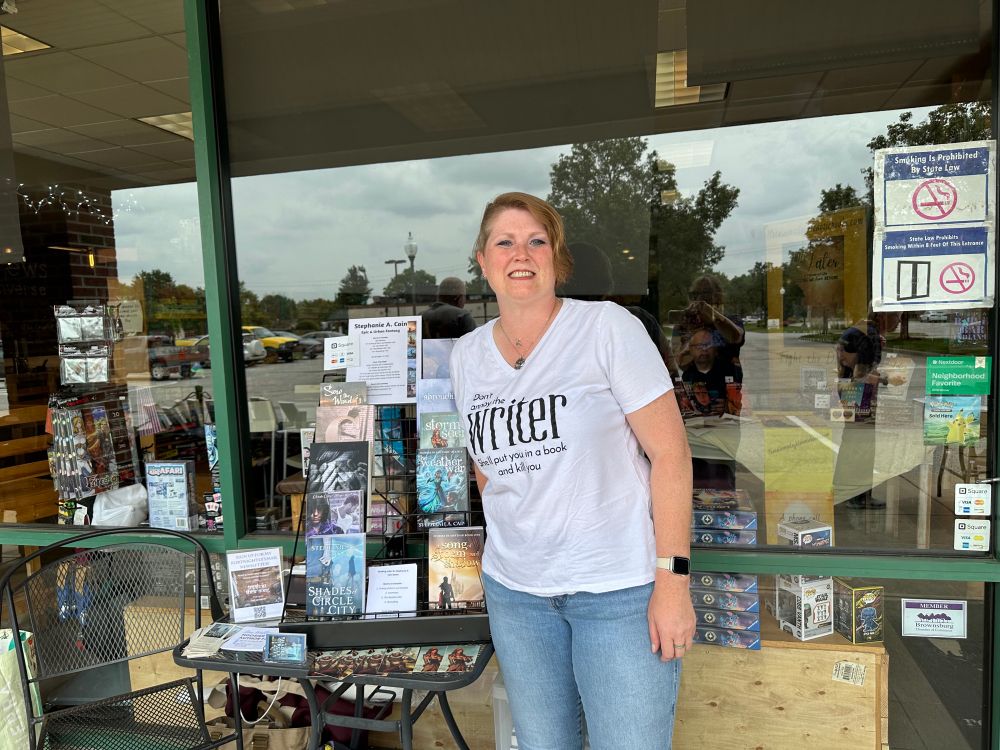Author Stephanie A. Cain, a red haired woman, stands in front of a book display at a bookstore. 