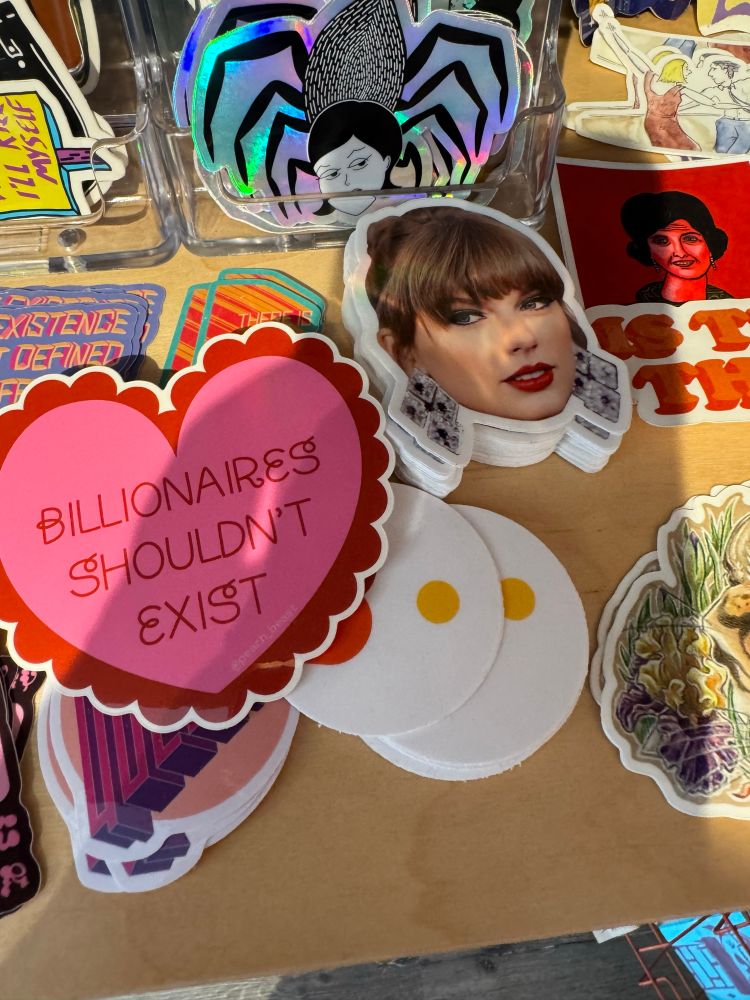 Array of stickers at indie bookshop. One says “billionaires should not exist”, the other is the face of billionaire Taylor Swift