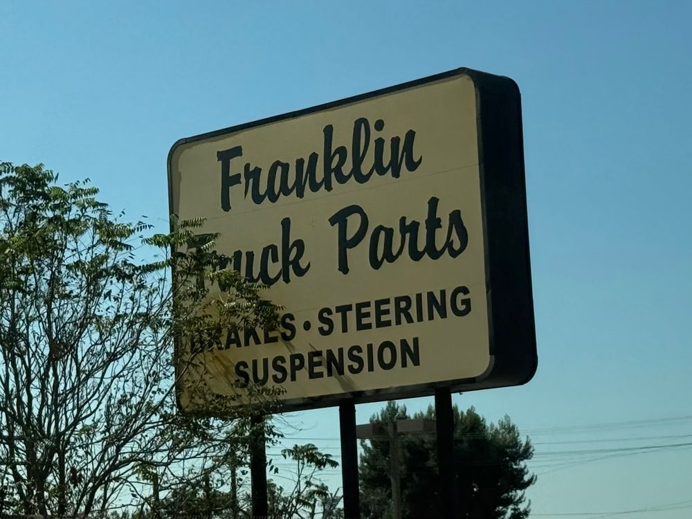 A road-side sign for “Franklin Truck Parts”. The sign is partially concealed by a tree, which makes the word “Truck” look like the word “Fuck” due to the choice of typeface.
