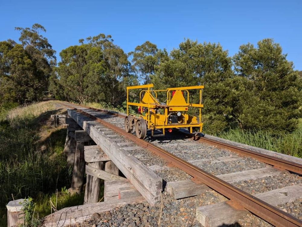 A yellow railway trolley sits atop a bridge on the track. The sky is blue and there's lots of trees in the background 