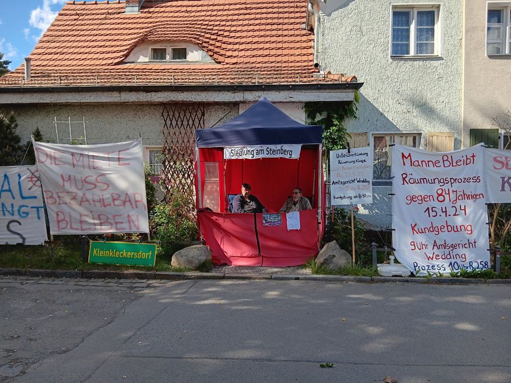 Man sieht einen roten Pavillon, darunter sitzen zwei Personen. Drum herum hängen viele Transparente.