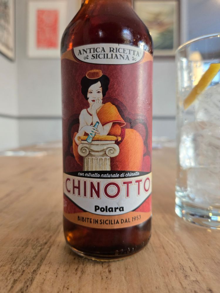 Eine Flasche Chinotto auf dem Tisch. Ein italienisches bzw. sizilianisches Erfrischungsgetränk mit etwas herber Note. Abgebildet ist eine Frau, die sich auf eine Säule stützt.