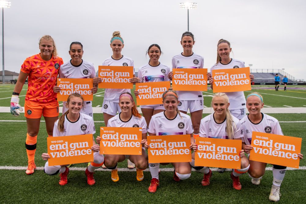 Sioux Falls City Football Club's inaugural home opener's starting eleven wearing all white kits and holding orange signs that read "we can end gun violence" in white text. The goalkeeper is in an all orange kit.