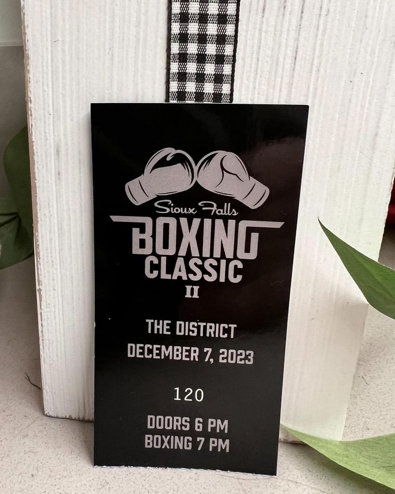 A black sign that reads “Sioux Falls Boxing Classic II. The District. December 7, 2013. Doors 6PM. Boxing 7PM.” in white text. At the top is white boxing gloves.