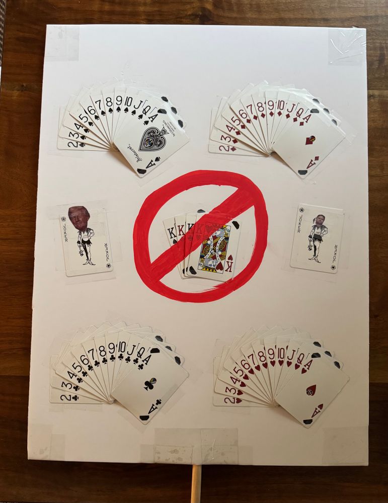 Protest sign of a deck of cards fanned out by suit. The Kings are in a separate pile with a no symbol drawn in red over the top of them to signify No Kings. The joker cards are also displayed with a picture of Trump and Vance’s head imposed over the jokers heads. 
