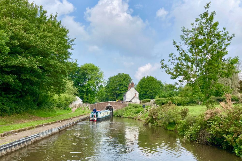 A view of the canal looking towards the bottom gates of a narrow lock. A narrow boat is getting ready to enter the lock, under the brick bridge that covers the entrance. The summer sun is shining and the trees and foliage  are in full bloom. A white cottage sits next to the lock.