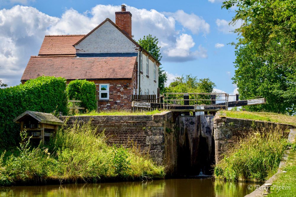 The picture shows the lower entrance to a canal lock, the lock gates are closed. On the left sits a large lock cottage. The lock is surrounded by grass and trees and the sky is blue, with some fluffy white clouds. The summer sun is shining and everything is bright and vivid.