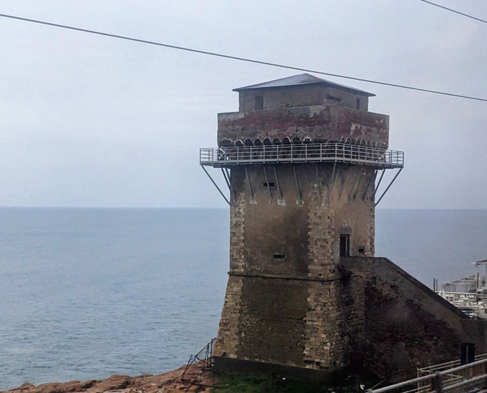 View out a train window looking towards a blueish grey sea under a grey sky; just right of center is an old watch tower built of cut stone and brick with a parapet above.