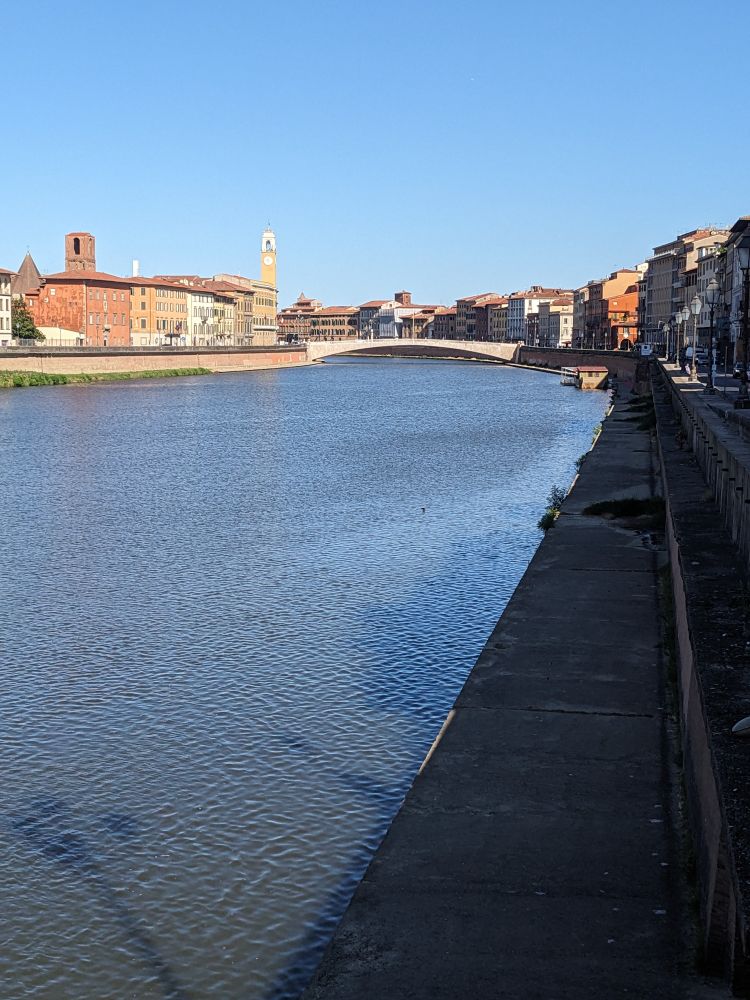 Photo of the Arno River at Pisa looking west, downstream, towards the Ponte di Mezzo, in early-mid morning light, the right bank in shadow, the left bank with its old, reddish ochre buildings in sun.