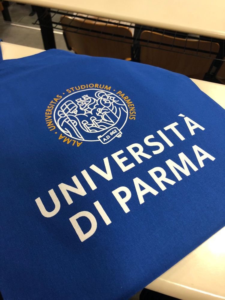 A blue tote bag laying on a table. The tote bag has the words "UNIVERSITA DI PARMA" on the front in while lettering. There is also a logo with the Latin phrase "ALMA . UNIVERSITAS . STUDIORUM . PERMENSIS".
