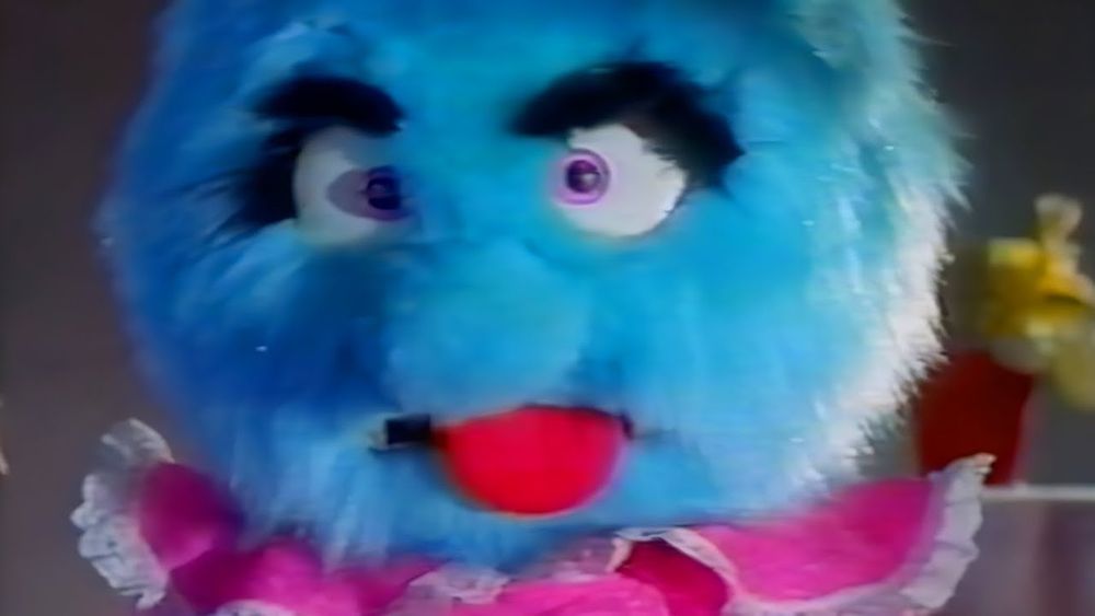 Blue puppet in a pink dress sticking out its tung. 