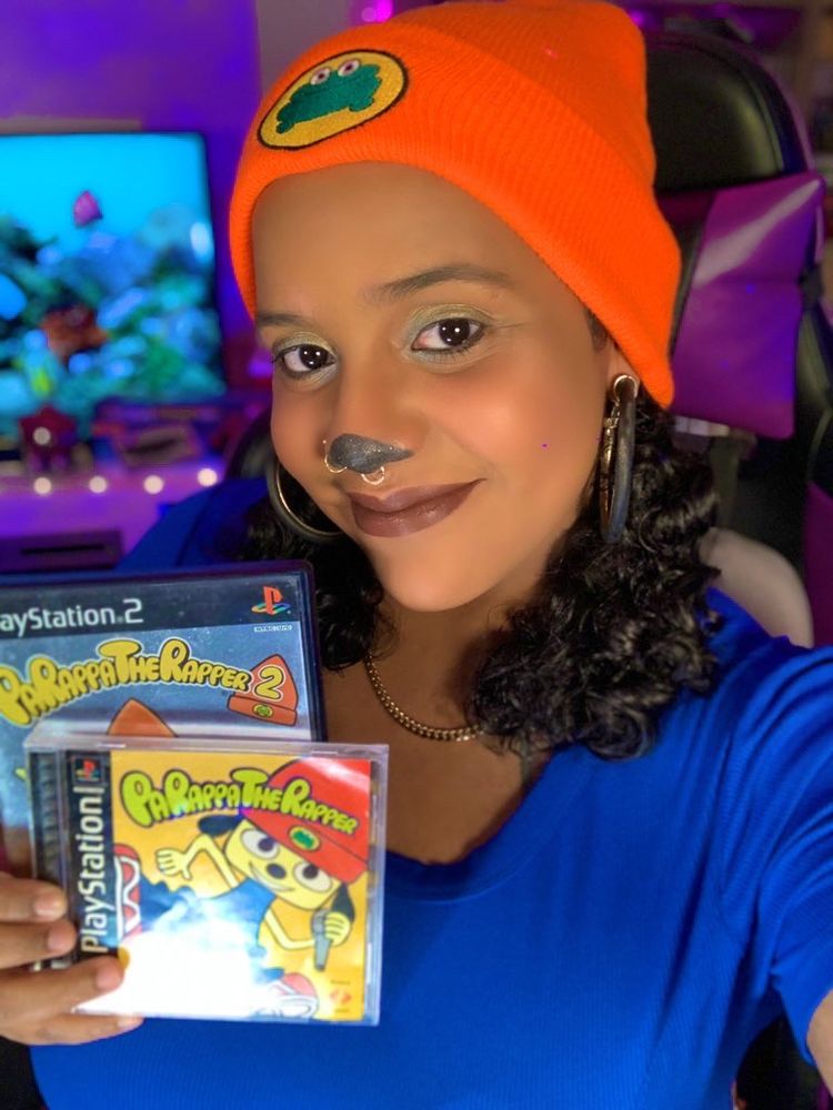 Zeph takes a selfie wearing a PaRappa the Rapper cosplay. She wears a solid blue top, an orange beanie with a frog, and her hair is in two small pigtails. She holds both PaRappa the Rapper for PS1 and PaRappa the Rapper 2 for PS2 in her right hand.