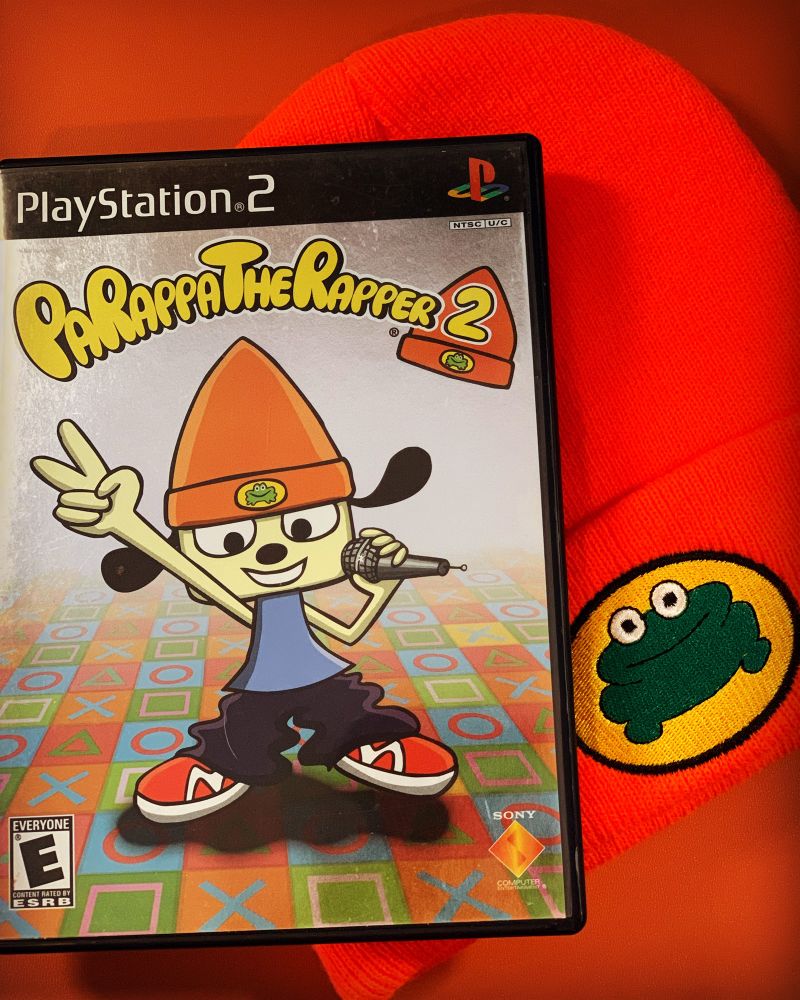 The video game PaRappa the Rapper 2 for PS2 sits on a burnt orange background. The cover art features an animated dog character holding a microphone rapping. Next to the video game is an orange beanie with a frog in the middle.