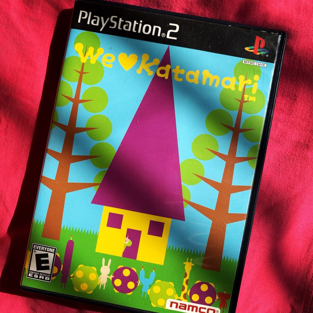 The video game “We Love Katamari” for PS2 lay on top of a pink background. The cover is very colorful and has a yellow and purple house on it.