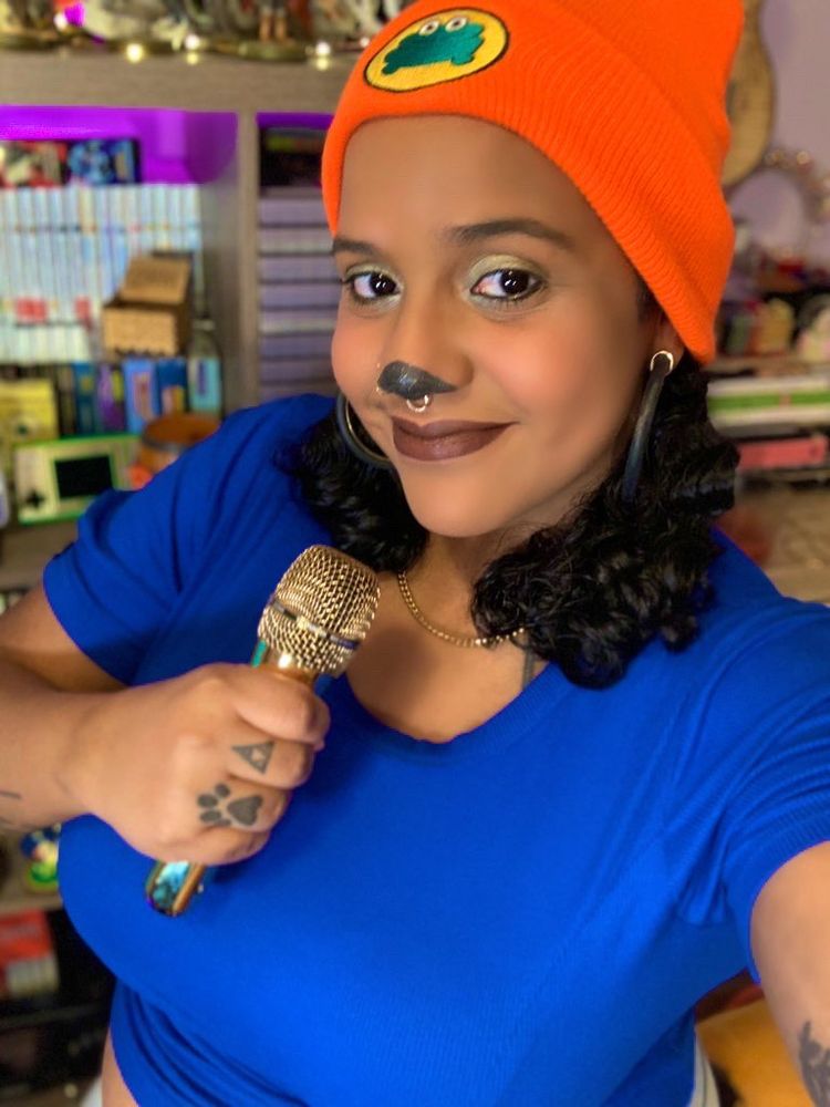 Zeph takes a selfie wearing a PaRappa the Rapper cosplay. She wears a solid blue top, an orange beanie with a frog, holds a gold mic and her hair is in two small pigtails.