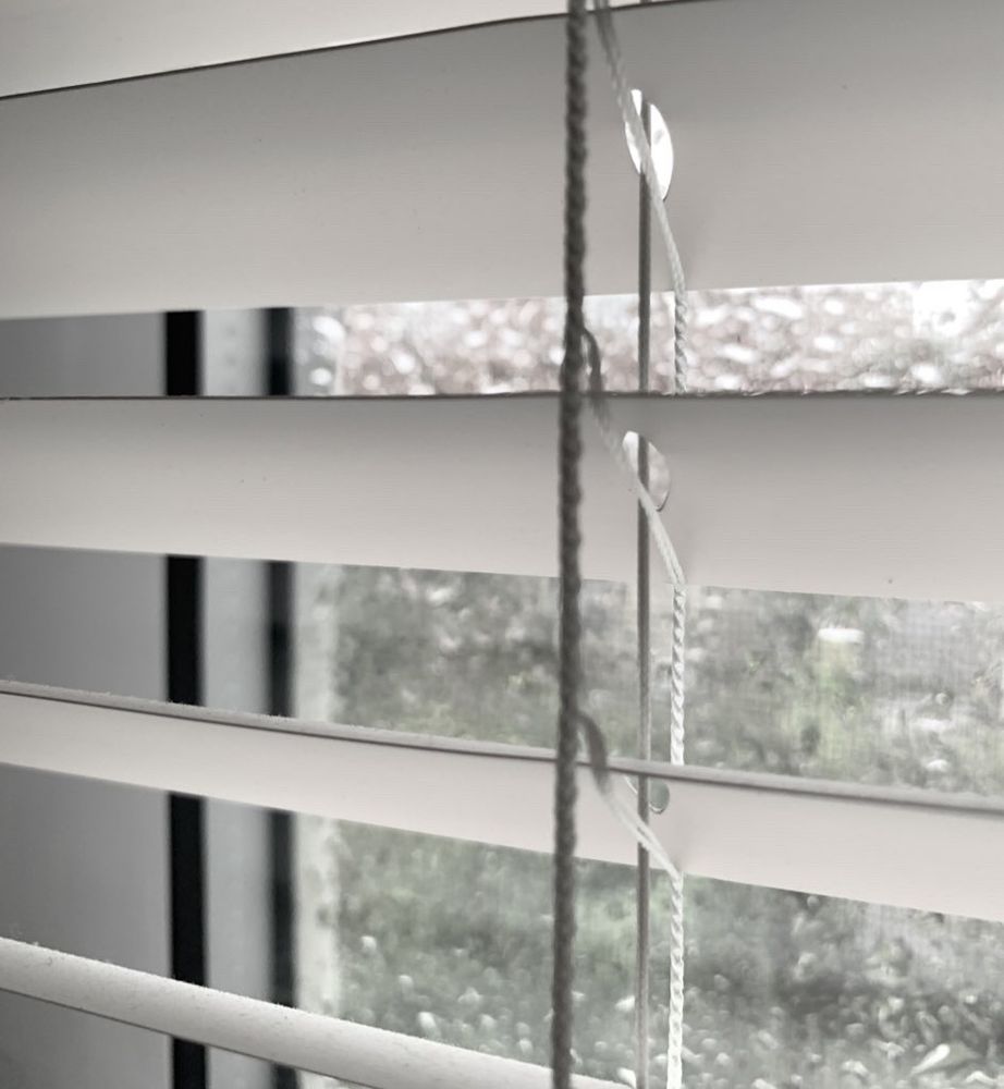 Up close picture of white window blinds slightly opened and showing a window full of rain drops.