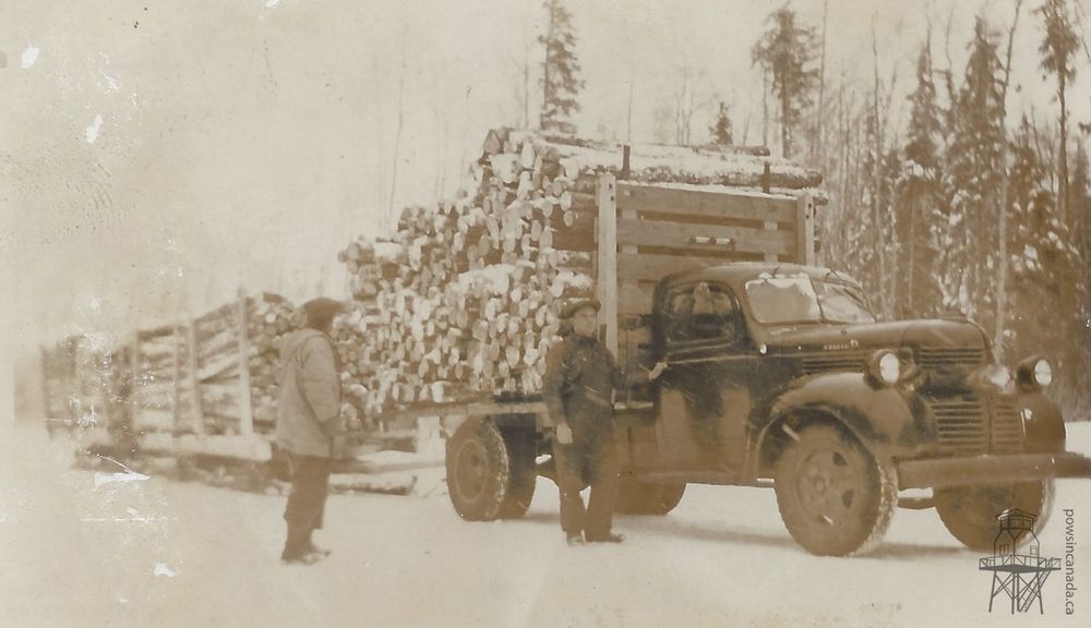 Black and white photo of two men standing next to a truck hauling loads of pulpwood.