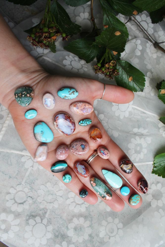 an image of an outstretched hand in front of a background of white floral fabric and greenery. On the hand there are a number of different cabochons of different sizes and shapes, including Kingman Turquoise, Mexican fire opal, and other precious stones. They will be turned into handmade jewelry by Andisa Studio owner Isabelle Anderson
