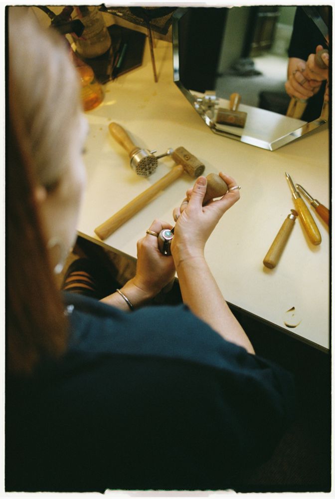A film photo over the shoulder of a redhead woman wearing a black shirt. she is using jewelry making tools to finish a sterling silver ring set with a turquoise precious stone. Made by Andisa Studio Isabelle Anderson photo by Charlotte J Mack