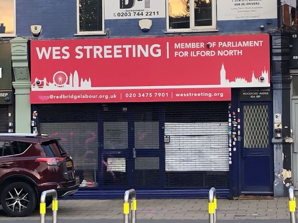 A photograph of the front of Wes Streeting's office in Ilford. there is pink paint on the window, reading CHILD KILLER, although a car is parked in front of it, and white paper on one side barely hiding a puncture in the glass. Wes' name is also covered in pink paint on the email at the bottom of the red banner-style sign